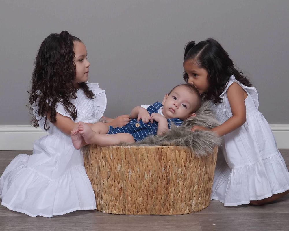 2 girls with white dress holding a boy in blue laying on a wood table with grey background wall. 